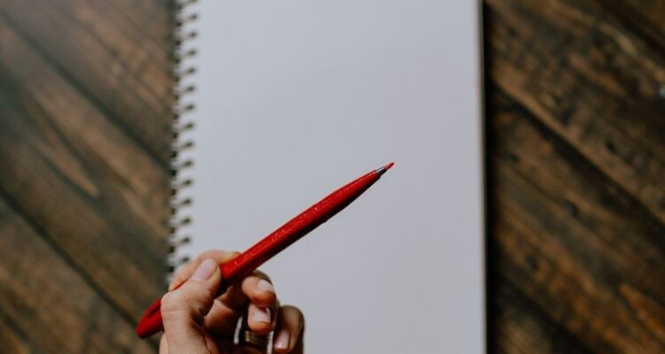 Hand hält einen roten Stift über einem blanken Spiralnotizbuch
